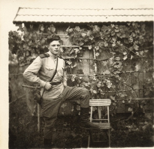 Mężczyzna w sowieckim mundurze pozuje do zdjęcia na tle wiejskiej chaty, lewą nogę trzyma opartą na krześle