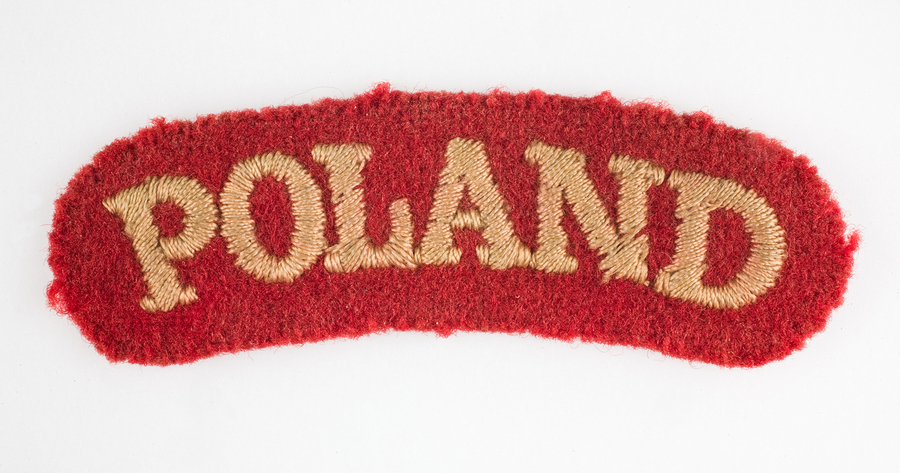 Noszona przez por. Władysława Jaksana odznaka naramienna 2 Korpusu Polskiego – Poland. Napis wyhaftowany nicią koloru łososiowego na materiale koloru czerwonego.