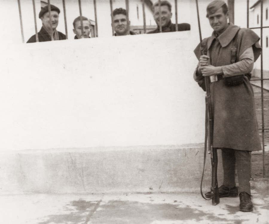 Zdjęcie 021. Czterech więźniów stojących za murem i spoglądających przez kraty. Trzeci od lewej por. Franciszek Cieplik. Przed murem, niedaleko furty z metalowych prętów stoi uzbrojony wartownik. Prawdopodobnie jest to hiszpański obóz Miranda de Ebro. Więźniowie i strażnik w ubiorach zimowych. Zdjęcie zostało wykonane najprawdopodobniej w okresie od grudnia 1941 do marca 1942 roku.