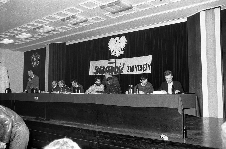 Zdjęcie 027. Lech Wałęsa i jego współpracownicy przy stole prezydialnym w Sali BHP Stoczni Gdańskiej im. Lenina w dniu spotkania z kandydatami do sejmu i senatu 29 kwietnia 1989 r. Od lewej: Andrzej Wielowieyski (przemawia), Bronisław Geremek, Lech Wałęsa, Henryk Wujec, Władysław Frasyniuk, Andrzej Stelmachowski (stoi zasłonięty przez Janusza Pałubickiego), Alojzy Pietrzyk i Piotr Nowina-Konopka. Fot. Igor Witowicz.
