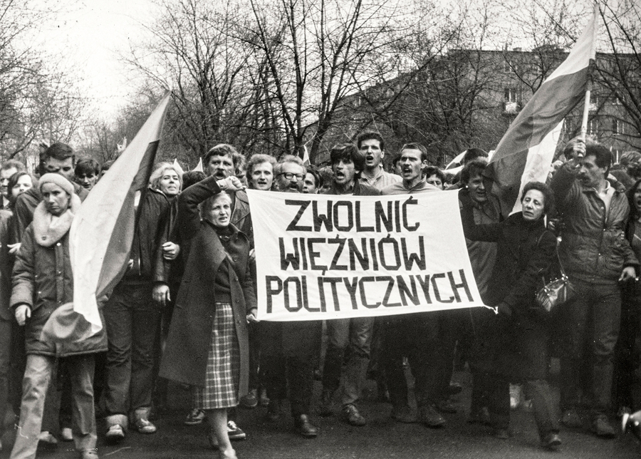Zdjęcie 015. Sympatycy i działacze NSZZ „Solidarność” idą w pochodzie ulicą Stołeczną w Warszawie 1 maja 1985 r. Na zdjęciu widoczni są działacze NSZZ „Solidarność”, m.in. Seweryn Jaworski, Henryk Wujec i Maciej Jankowski. Przed nimi idą kobiety trzymające transparent z hasłem: „ZWOLNIĆ WIĘŹNIÓW POLITYCZNYCH”.